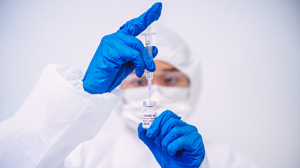 Doctor in protective gloves & workwear filling injection syringe with COVID-19 vaccine